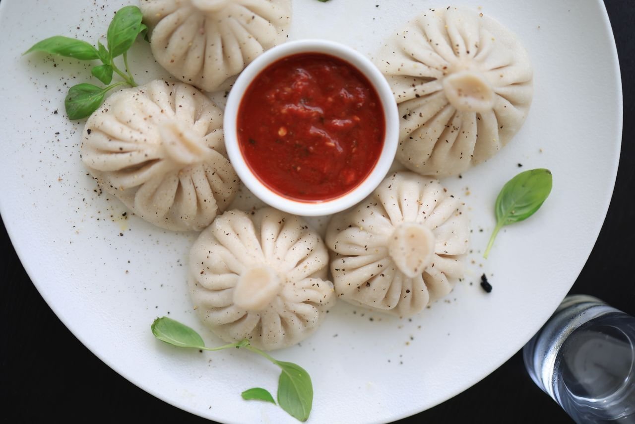 gallery-1 Five steamed dumplings with red sauce and basil on a white plate, food close-up.